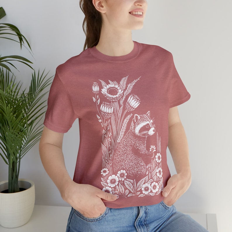 May include: A maroon t-shirt with a white graphic of a raccoon sitting in a field of flowers. The raccoon is holding a flower in its paw.