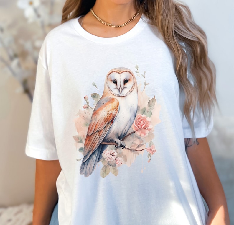 K&ouml;nnte beinhalten: Wei&szlig;es T-Shirt mit einer Aquarell-Illustration einer Schleiereule, die auf einem Ast mit rosa Bl&uuml;ten sitzt.