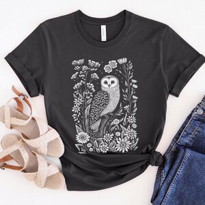 May include: A black t-shirt with a white print of an owl surrounded by flowers. The owl is perched on a branch and has its wings spread out. The flowers are in bloom and have white petals.