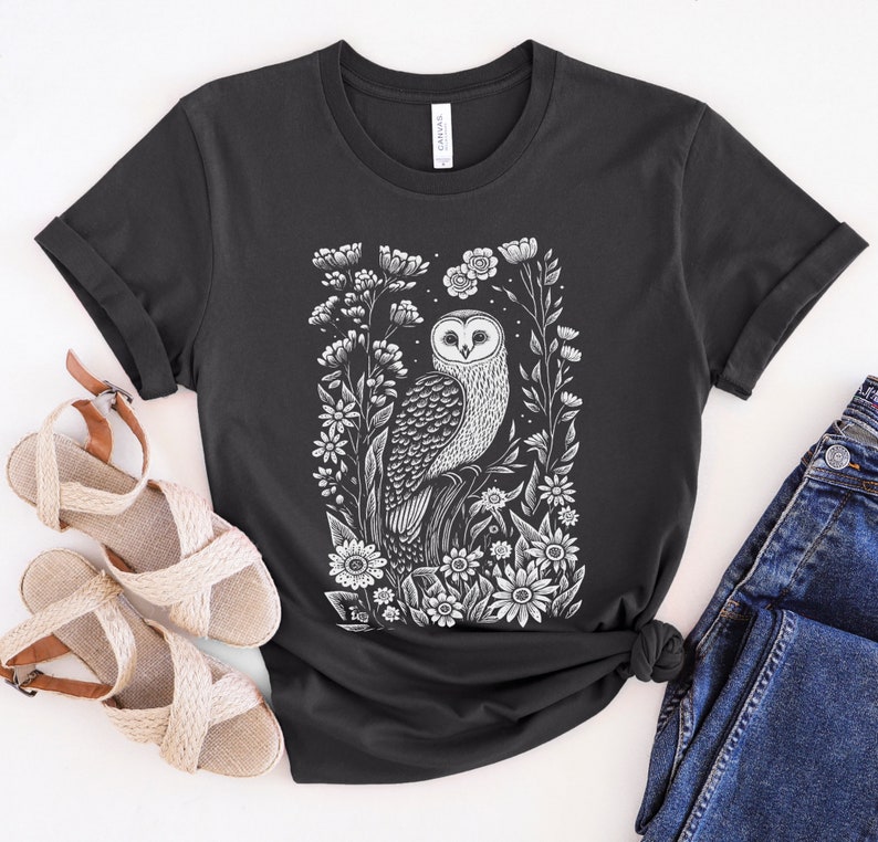 May include: A black t-shirt with a white print of an owl surrounded by flowers. The owl is perched on a branch and has its wings spread out. The flowers are in bloom and have white petals.