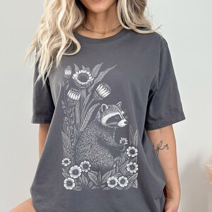May include: A charcoal gray t-shirt with a white graphic of a raccoon surrounded by flowers. The raccoon is looking to the right and holding a flower in its paw.