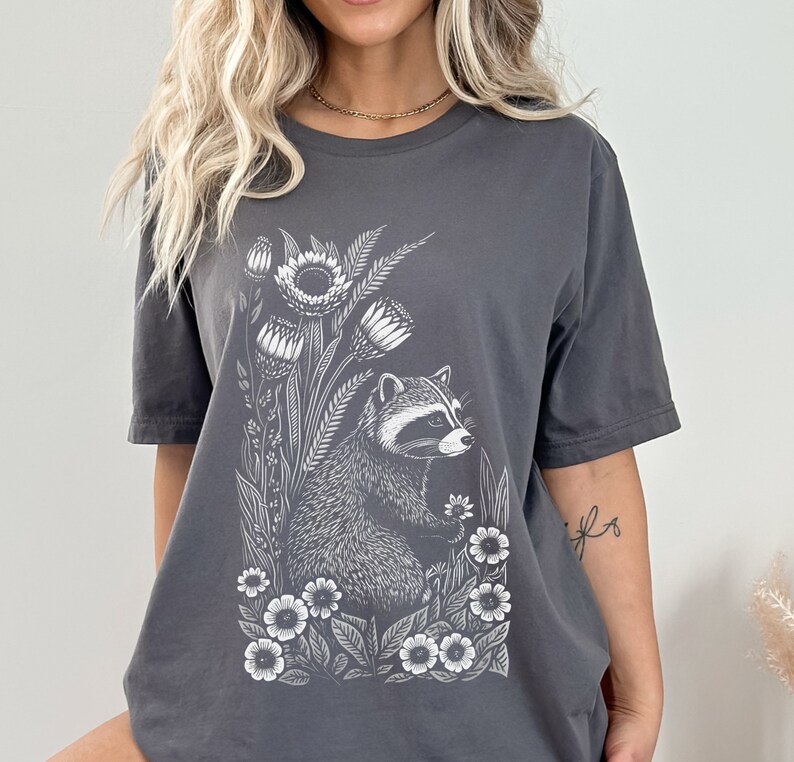 May include: A charcoal gray t-shirt with a white graphic of a raccoon surrounded by flowers. The raccoon is looking to the right and holding a flower in its paw.