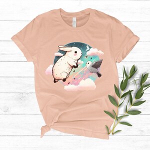 May include: A peach-colored t-shirt with a graphic of a white rabbit and a gray bird flying in a colorful sky with clouds. The graphic is surrounded by a circle.
