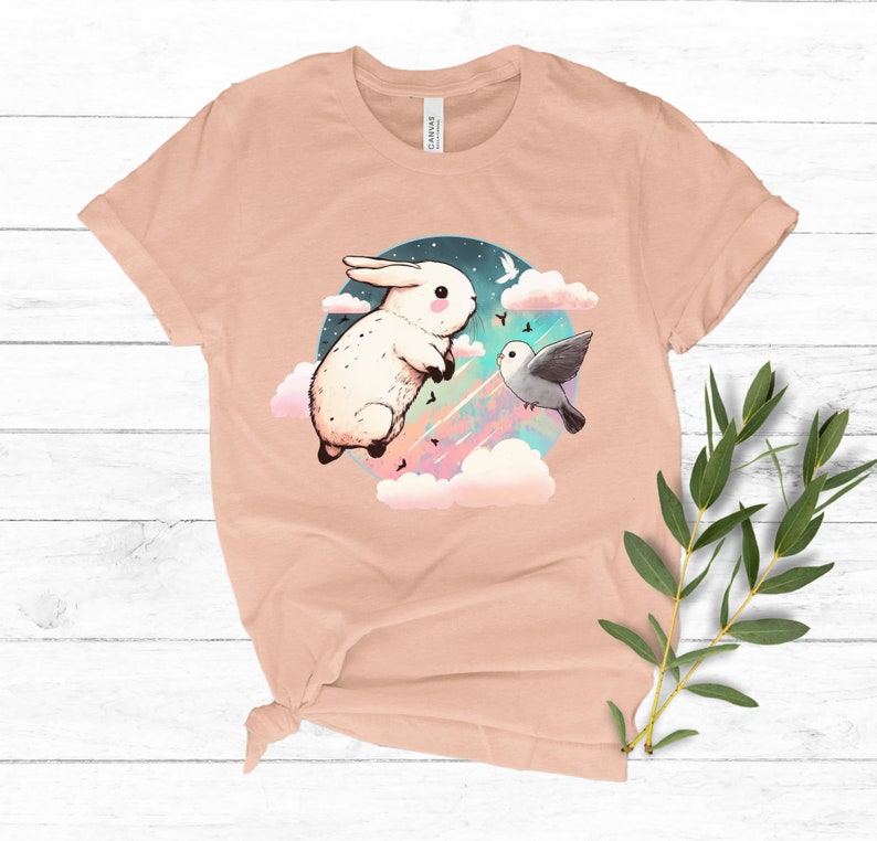 May include: A peach-colored t-shirt with a graphic of a white rabbit and a gray bird flying in a colorful sky with clouds. The graphic is surrounded by a circle.