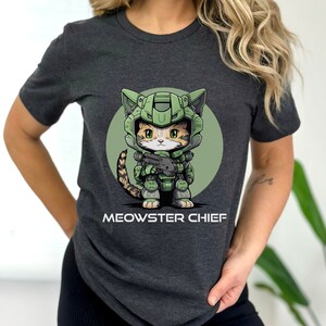 Puede incluir: Camiseta gris oscuro con un gato de dibujos animados con armadura verde, sosteniendo un arma, sobre un c&iacute;rculo verde. El texto "MEOWSTER CHIEF" est&aacute; impreso debajo. La camiseta es de manga corta.