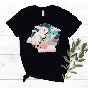 May include: Black t-shirt with a white rabbit and a bird flying in a circle of clouds against a dark blue background with stars and a rainbow.