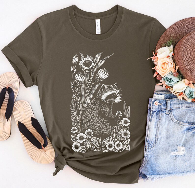 May include: A dark green t-shirt with a white graphic of a raccoon surrounded by flowers. The raccoon is looking to the right and holding a flower in its paw.