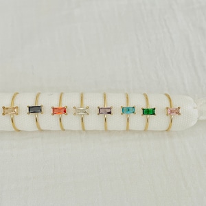 Könnte beinhalten: Ein Satz von zehn goldenen Ringen mit bunten rechteckigen Edelsteinen. Die Ringe sind in einer Reihe auf einer weißen Oberfläche angeordnet.