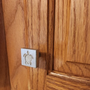 May include: A silver square cabinet knob with a raised, engraved image of a sea turtle.