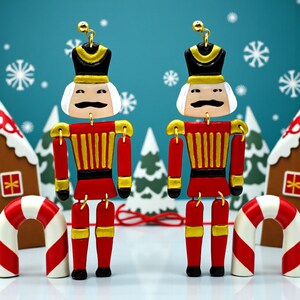 May include: Two red and gold nutcracker ornaments with black hats and white hair, set against a winter scene with snowflakes, gingerbread houses, and candy canes. The nutcrackers have black moustaches and are connected with gold accents.