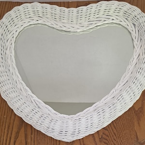 May include: A heart-shaped wall mirror featuring a white woven wicker frame. The reflective glass surface is centered within the braided border, which follows the curved outline of the heart shape to create a decorative and functional home accessory.