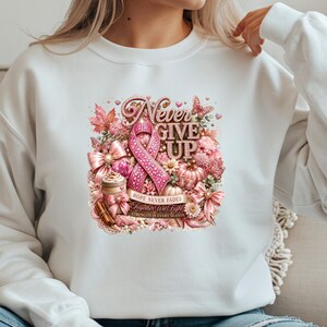 May include: A white crewneck sweatshirt featuring a central graphic design with a pink awareness ribbon, the text "Never Give Up," "Hope Never Fades," "Together We Fight," and "Strength In Every Season," surrounded by pink flowers, bows, and pumpkins.