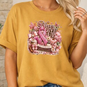 May include: A mustard yellow t-shirt featuring a graphic print with a pink ribbon, flowers, pumpkins, and bows. The text reads Never Give Up, Hope Never Fades, Together We Fight, and Strength In Every Season in a decorative, glittery font style.