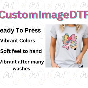 May include: A light gray t-shirt featuring a graphic of a large, multi-colored bow with a red heart in the center, positioned above a pencil. The text 100 days of school is printed in a stylized font across the center of the graphic design.