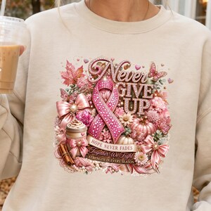 May include: A beige crewneck sweatshirt featuring a graphic print of a pink breast cancer awareness ribbon, a coffee cup, pumpkins, bows, and autumn leaves. The text reads: Never Give Up, Hope Never Fades, Together We Fight, Strength In Every Season.