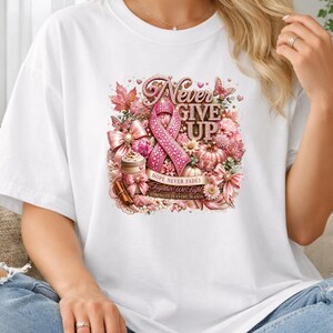 May include: A white short-sleeve t-shirt featuring a central graphic with a pink ribbon, autumn leaves, pumpkins, and a coffee cup. The text reads Never Give Up, Hope Never Fades, Together We Fight, and Strength In Every Season in pink and gold.