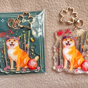 Könnte beinhalten: Zwei transparente Acryl-Schlüsselanhänger mit einem Cartoon-Shiba-Inu-Hund, der ein rotes Stirnband mit den japanischen Schriftzeichen "樂天" (Rakuten) trägt. Der Hund sitzt vor einem Bambuswald und einer roten Daruma-Puppe. Die Schlüsselanhänger sind an goldfarbenen Metallringen befestigt.