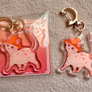 Può includere: Due ciondoli portachiavi in ​​acrilico trasparente con un gatto dei cartoni animati che indossa un cappello arancione. I ciondoli sono confezionati in sacchetti di plastica con una carta bianca su cui è scritto "My Kittens" e "Acrylic charms by Sara Cervasi".