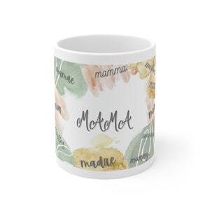 May include: White ceramic mug with a watercolor design of green and peach colored abstract shapes. The words "mamma", "mummy", "mae", "madre", "mom", and "mother" are written around the mug. The text "McA McA" is written in the center of the mug.