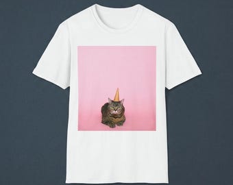 Puede incluir: Una camiseta blanca de manga corta con un estampado gráfico centrado de un gato atigrado marrón que lleva un gorro de fiesta cónico de color tostado. El gato está situado sobre un fondo rosa claro sólido, enmarcado en un cuadrado en la camiseta.