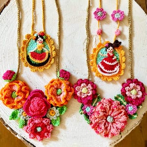Op de afbeelding: Drie gehaakte kettingen met bloem- en poppenversieringen. De kettingen zijn gemaakt van kleurrijk garen en bevatten verschillende bloemen, waaronder roze, rood en oranje. Elke ketting heeft een kleine pop in het midden, die een kleurrijke jurk draagt.