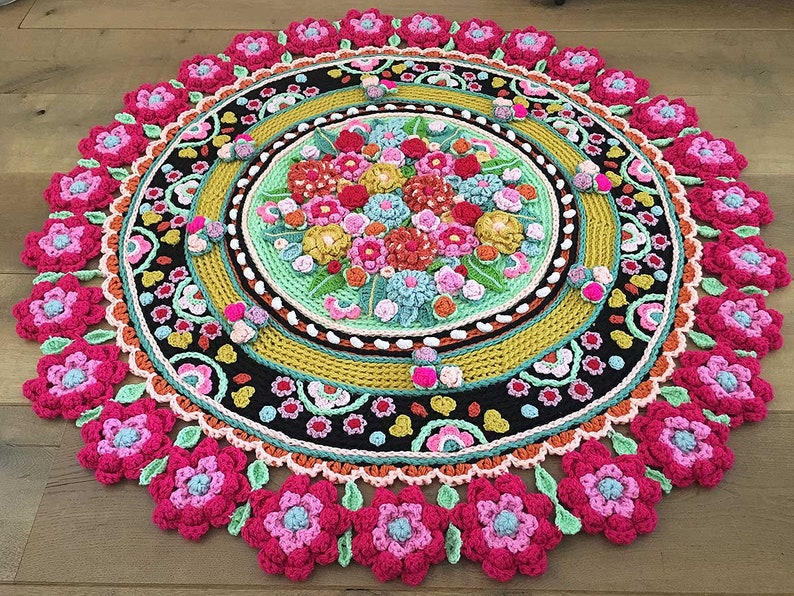 Peut inclure: Un tapis rond en crochet de style mandala avec un motif floral dans des tons de rose, vert, jaune et orange. Le tapis a un centre noir avec une bordure multicolore.