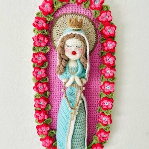 Op de afbeelding: Gehaakte afbeelding van de Maagd Maria, die een blauwe en groene jurk draagt met een gouden kroon en een rozenkrans vasthoudt. De figuur is omgeven door een roze gehaakte ovaal met een rand van rode bloemen.