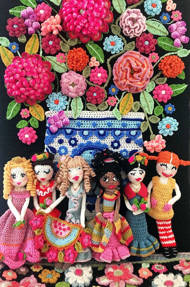 May include: Seven crocheted dolls in various colors and styles stand in front of a crocheted flower arrangement in a blue and white vase. The dolls are wearing dresses and skirts with colorful details. The flowers are in shades of pink, red, orange, yellow, and blue.