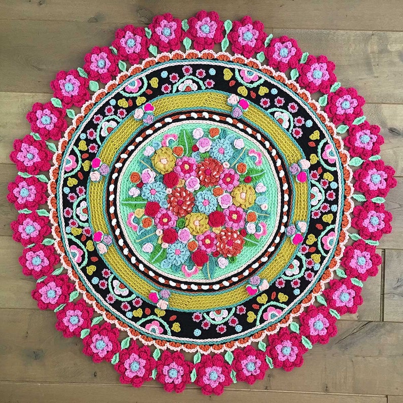 Peut inclure: Un tapis rond en crochet de style mandala avec un motif floral dans des tons de rose, jaune, orange et vert. Le tapis a un centre noir avec une bordure multicolore.