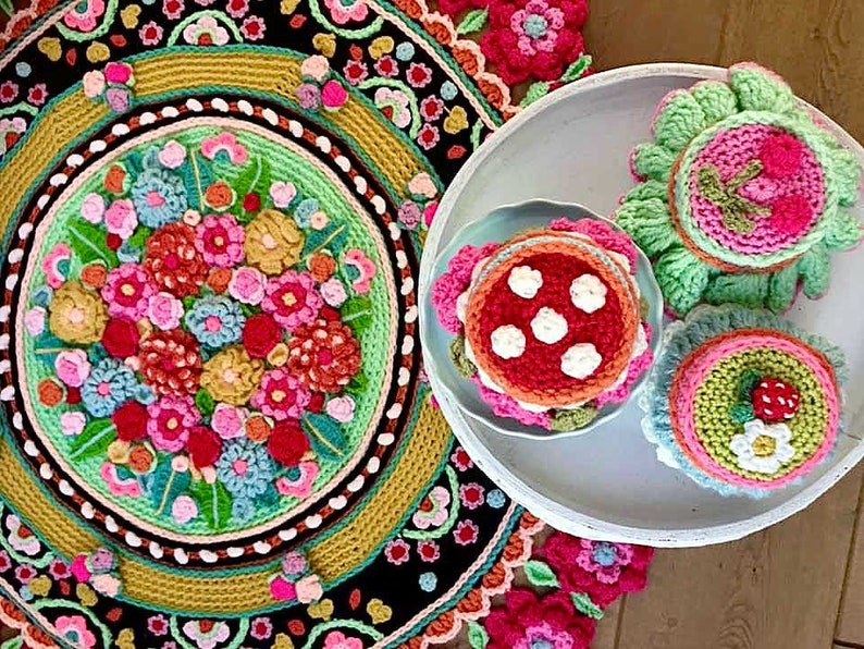 Peut inclure: Un tapis en crochet en forme de mandala avec un motif floral dans des tons de rose, rouge, vert et jaune. Trois g&acirc;teaux en crochet avec un gla&ccedil;age rose, vert et blanc sont pr&eacute;sent&eacute;s sur un plateau blanc.