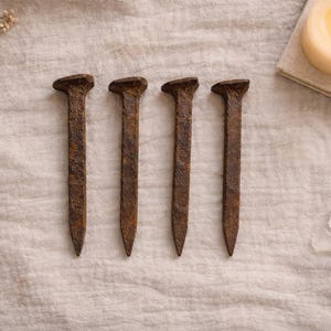 May include: Four rusty iron nails with square heads are arranged on a textured, off-white surface. A small bowl of granules, a lit candle, and a few clear crystals are also present, suggesting a rustic or spiritual theme.