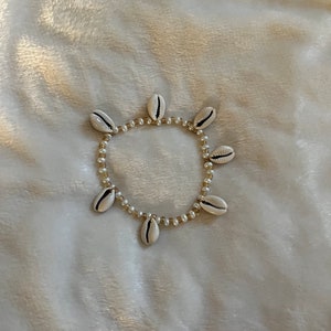 May include: A delicate bracelet featuring a circular design of small, white seashells with black markings. The shells are interspersed with small, round, off-white beads, creating a natural, beachy aesthetic. The bracelet rests on a soft, white, textured surface.