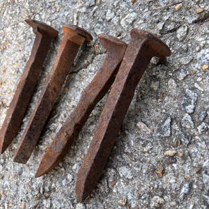 Puede incluir: Cuatro clavos de ferrocarril oxidados, probablemente utilizados para la construcción de vías, están dispuestos sobre una superficie de hormigón gris rugosa. Los clavos son de metal y tienen un extremo puntiagudo y una cabeza plana.