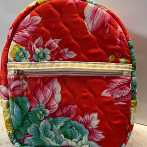 Peut inclure: Un sac à dos rouge matelassé avec un motif floral rose, vert et blanc. Le sac à dos a une fermeture éclair à carreaux jaunes et blancs et un petit charme. Le sac mesure environ 30 cm de haut et 25 cm de large.