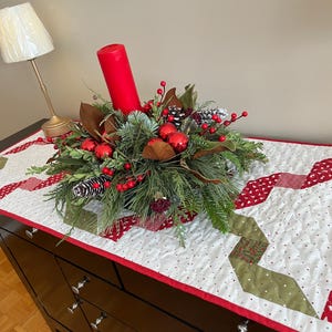 May include: A Christmas centerpiece featuring a red candle, red baubles, pine cones, and foliage. The arrangement is on a white quilted table runner with red and green details, placed on a dark brown dresser.