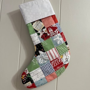 Peut inclure: Une chaussette de Noël en patchwork avec un revers blanc et un motif vert, rouge et bleu. La chaussette présente une variété de motifs, notamment des flocons de neige, des cannes en sucre et un bonhomme de neige. Le texte "Candy Canes" et "Nicholas Cakes" est visible sur la chaussette.