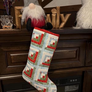 Peut inclure: Une chaussette de Noël matelassée avec un motif patchwork rouge, vert et bleu clair. La chaussette est ornée d'un motif de cabane en rondins et d'une bordure rouge. Elle est suspendue devant une cheminée.