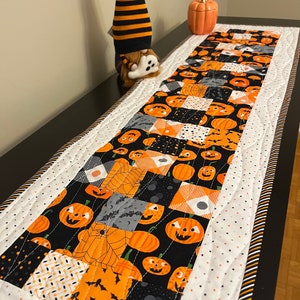 May include: A Halloween table runner with a patchwork design featuring pumpkins, bats, and spiderwebs on a black background. The runner is bordered with white fabric dotted with small stars and a striped edge. A decorative gnome and pumpkin are also present.