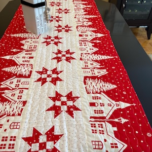 Puede incluir: Un camino de mesa navideño rojo y blanco con un patrón repetido de casas, árboles y estrellas. El centro presenta grandes estrellas rojas. Los bordes tienen un patrón de puntos. Una caja blanca con texto está sobre la mesa.