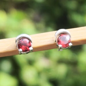 May include: Two small stud earrings with a silver setting, each holding a bright red gemstone. The earrings are presented on a light brown surface, set against a blurred green backdrop.
