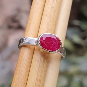 Puede incluir: Un anillo de plata con una piedra ovalada de color rojo brillante. El anillo está colocado sobre una superficie de madera.