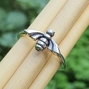 May include: A silver ring featuring a bee design. The bee is detailed with wings, a body, and legs. The ring is displayed on a wooden surface.