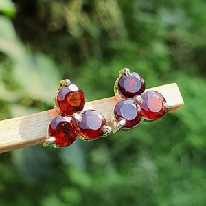 May include: A pair of stud earrings featuring six round, deep red gemstones set in silver. The earrings are arranged in a cluster design, with three stones on each earring. The background is a blurred green.
