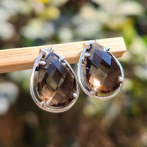 Puede incluir: Un par de pendientes en forma de lágrima con piedras de cuarzo ahumado facetadas engastadas en metal plateado. Los pendientes se muestran sobre un palito de madera sobre un fondo borroso verde y amarillo.