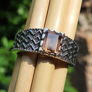 Könnte beinhalten: Ein silberner Ring mit geometrischem Muster und einem braunen Edelstein. Der Ring liegt auf einer Holzoberfläche.