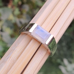 Puede incluir: Un anillo de plata con una piedra de luna rectangular engastada en el centro. El anillo está sobre una superficie de madera.