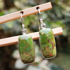 Puede incluir: Un par de pendientes rectangulares con una vibrante piedra verde y un borde de metal plateado. La piedra verde tiene un patrón único de vetas marrones y doradas. Los pendientes cuelgan de ganchos plateados.