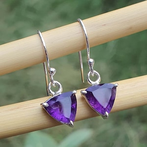 May include: Silver drop earrings with triangular, deep purple amethyst gemstones. The earrings hang from a simple hook design. The vibrant purple stones are displayed on a wooden dowel against a blurred green background. The earrings are a classic jewellery item.