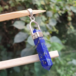 Pode incluir: Um pingente de prata com uma pedra preciosa de lapis-lazúli azul escuro em forma pontiaguda. O pingente está suspenso de uma fivela de prata.