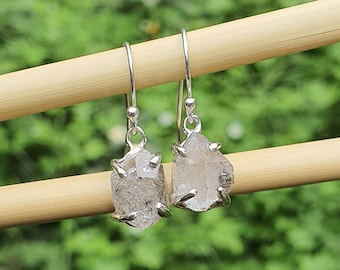 May include: A pair of silver earrings with clear, faceted, raw crystal stones hanging from the bottom. The earrings are hanging from a wooden stick.